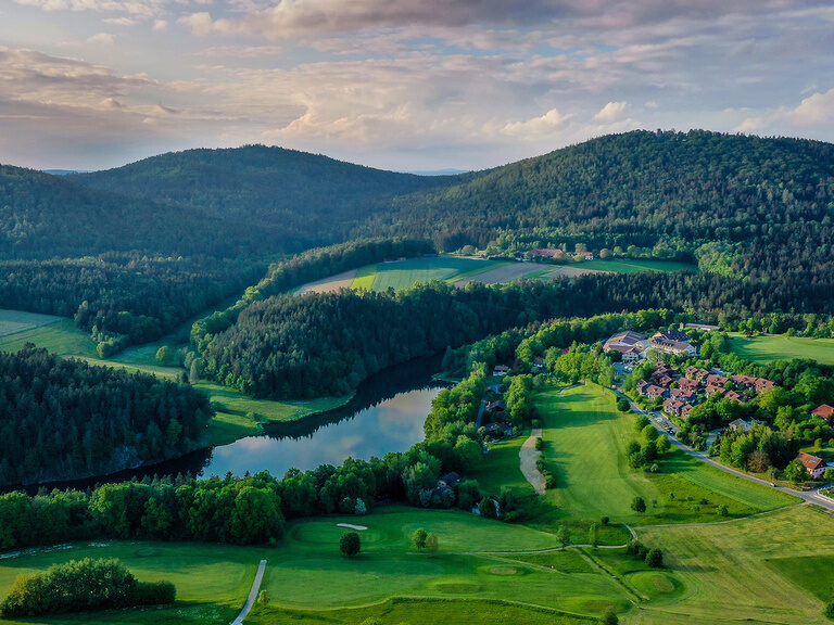 Das Hotel Wutzschleife eingebettet in grüne Hügellandschaft mit See und Wäldern.