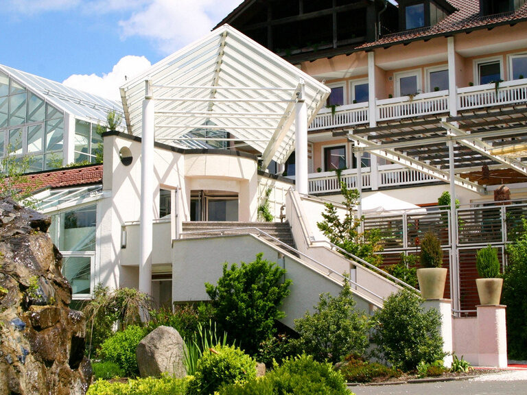 Hoteleingang mit Glasdach, Außentreppe, Natursteinbrunnen und üppiger Begrünung im Vordergrund.