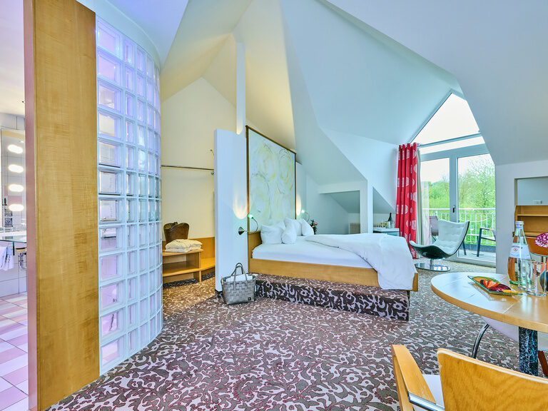 Helle Dachzimmer-Suite mit Glasblock-Badwand, freistehendem Bett, Balkon und Sitzbereich mit Tisch und Stuhl.