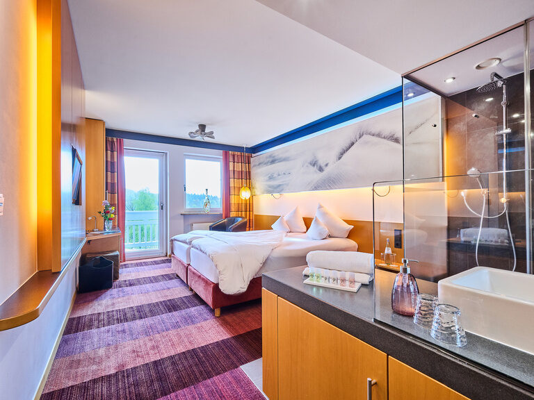 Hotelzimmer mit Panoramabett, offener Glasduschwand, lila Streifenteppich und Balkonblick.