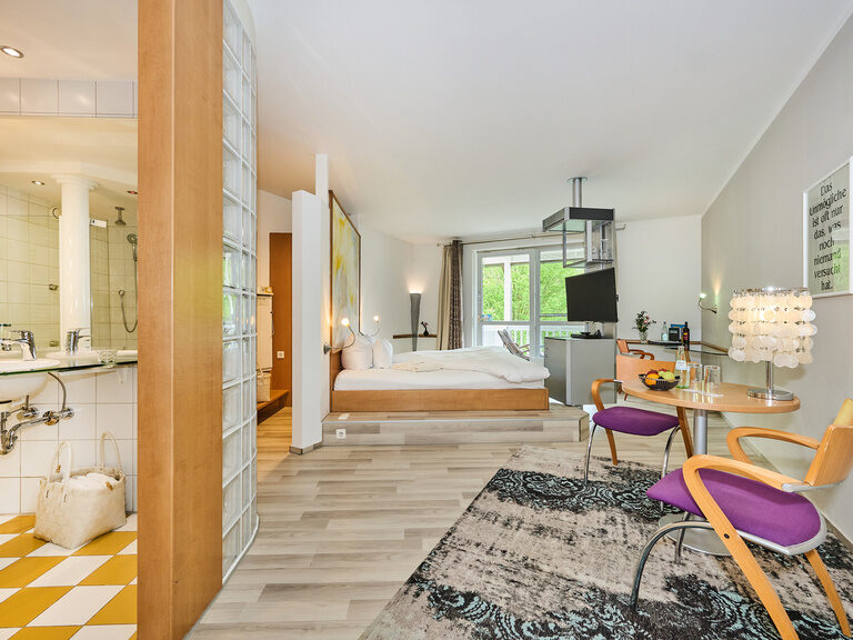 Offenes Hotelzimmer mit Glasbaustein-Duschwand, Panoramabett, lila Polsterstuhl und Teppich.