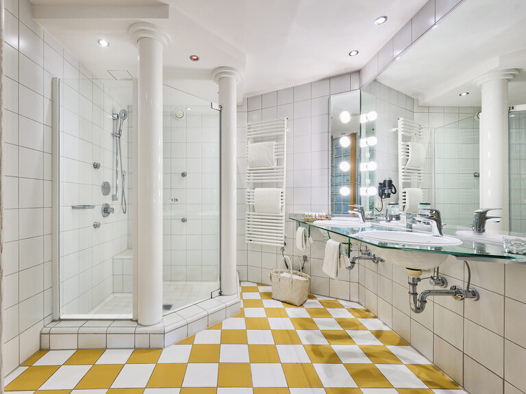 Dusche mit zwei Säulen, gelb-weiß kariertem Boden und Doppelwaschtisch auf Glasplatte.