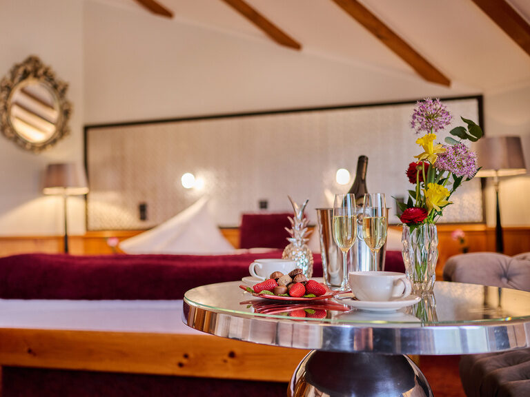 Hotelzimmer mit Blumen, Erdbeeren, Sektgläsern und gemütlichem Doppelbett im Hintergrund.