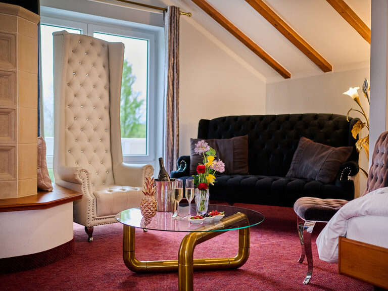 Dachschrägen-Sitzbereich mit weißem Ohrensessel, schwarzem Sofa, Glas-Couchtisch mit Blumen und Sekt.