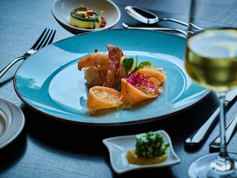 Gourmetgericht mit Garnelen, Gemüseröllchen und Kräutern auf blauer Keramiktellerplatte.