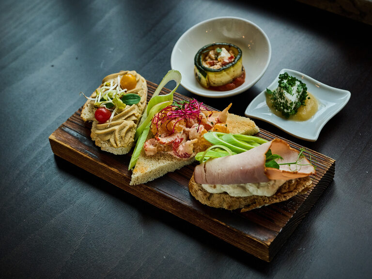 Drei kunstvoll belegte Gourmet-Häppchen auf Holzplatte mit dekorativen Kräutern angerichtet.