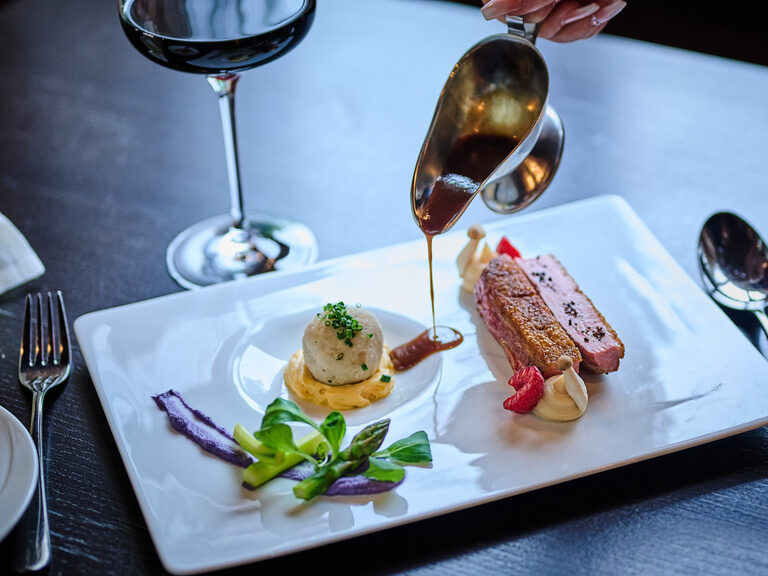 Fleischgericht mit Soße, Gemüse und Rotwein auf weißem Teller.