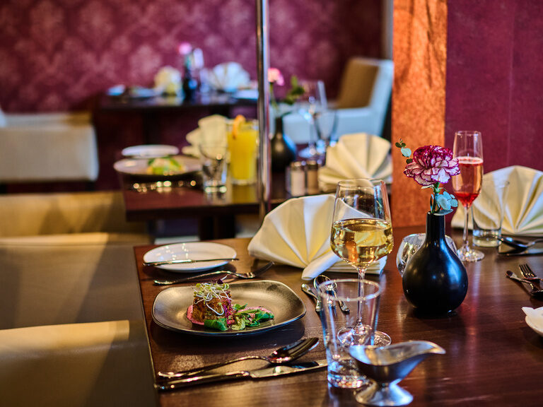 Edel gedeckter Zweiertisch mit Dessert, Wein und Blumen vor roter Wandverkleidung.