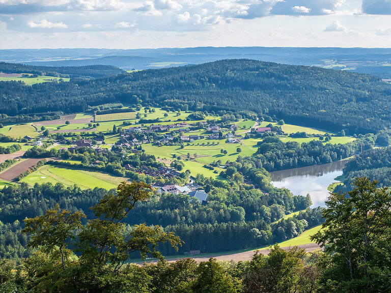 Weitläufige Hügellandschaft mit Wäldern, Dörfern und See bei Sonnenschein.