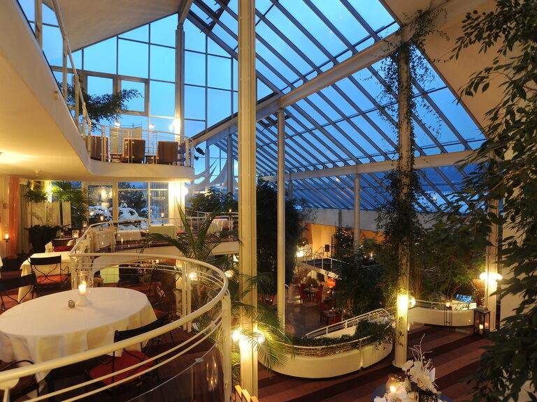 Wintergarten mit Kerzenlicht und mehreren Tischgruppen unter gläsernem Dach.
