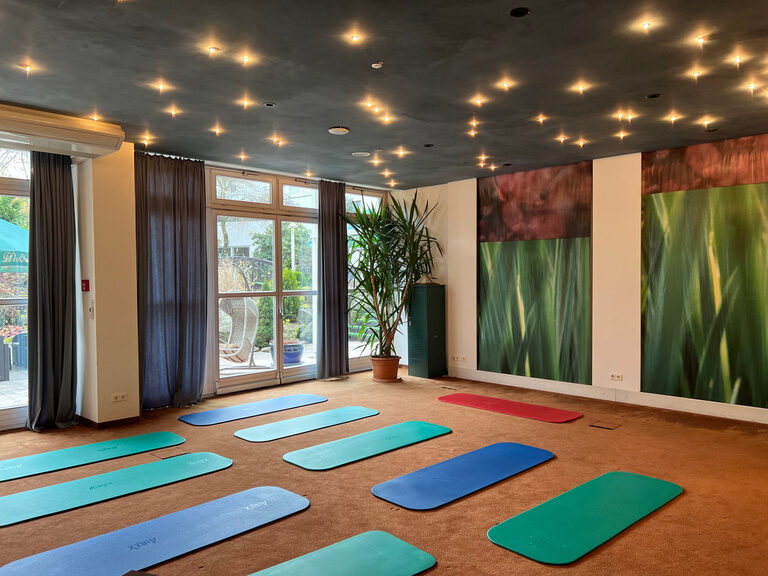 Heller Raum mit Yogamatten auf Teppichboden, Pflanzenmotiv-Wänden und Blick auf Terrasse.