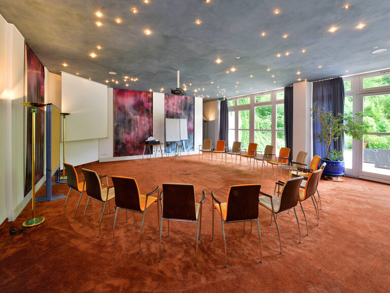 Stuhlkreis im Tagungsraum mit Panoramafenstern und Flipchart auf rötlichem Teppichboden.