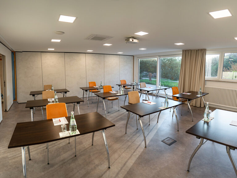 Seminarraum mit Mandala-Wandbild, Einzeltischen und Blick ins Grüne durch große Fenster.