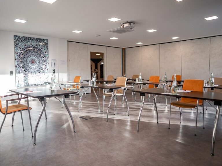 Seminarraum mit Einzeltischen, Fensterfront und Wand mit geometrischem Mandala-Muster.