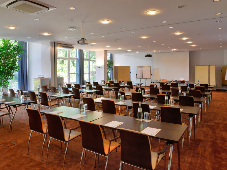 Großer Konferenzraum mit Reihenbestuhlung, Schreibmaterial und Tageslicht durch Fenster.