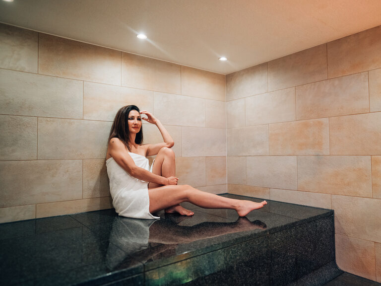 Frau sitzt entspannt in weißem Handtuch auf dunkler Steinbank im hellen Dampfbad.