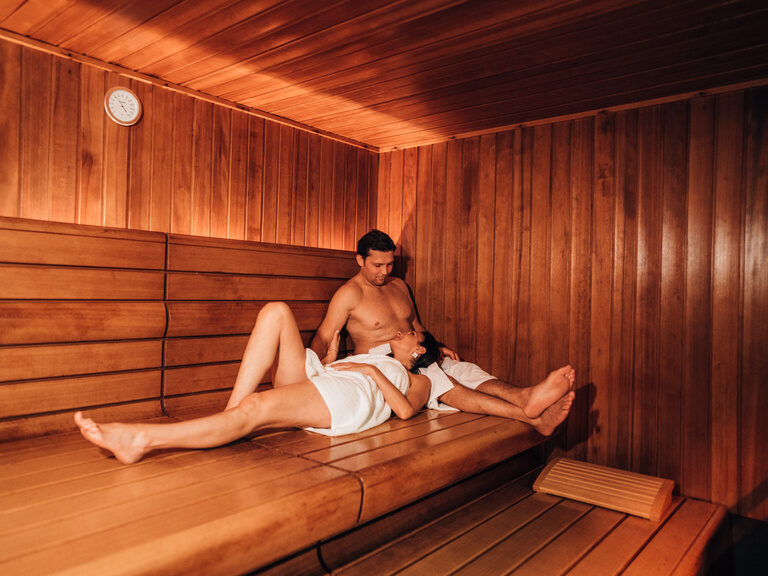 Paar entspannt in Sauna mit warmem Holzinterieur, gedimmtes Licht, intime Atmosphäre.