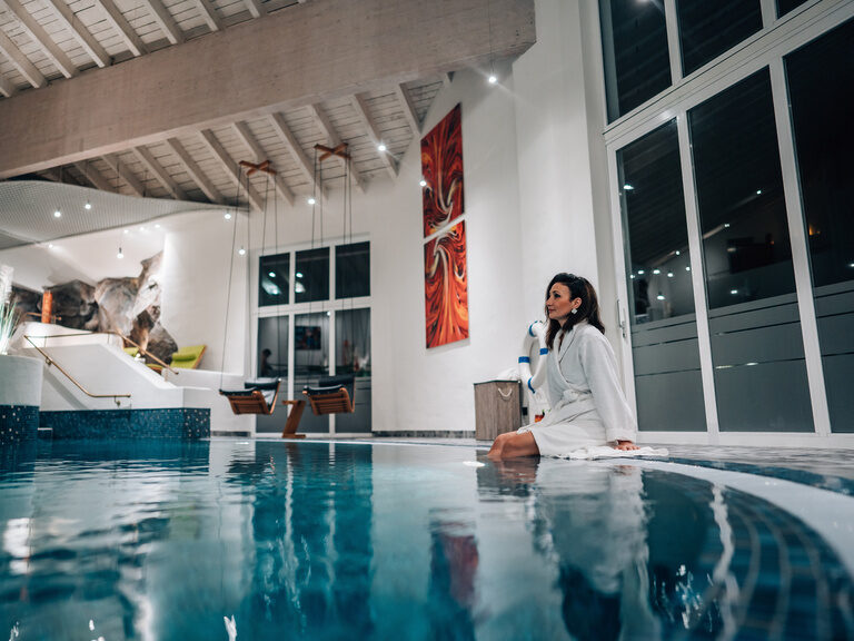 Frau im Bademantel sitzt am beleuchteten Innenpool in elegantem Spa-Bereich.