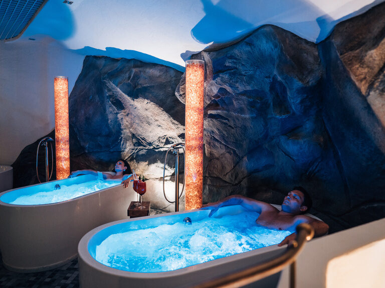 Entspannte Gäste in blauen, beleuchteten Badewannen vor Felswand mit Getränken auf Mini-Tisch.