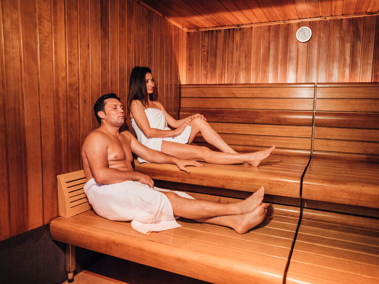 Paar in Holzsauna sitzt entspannt auf Bänken in zwei Etagen, Handtücher um den Körper.