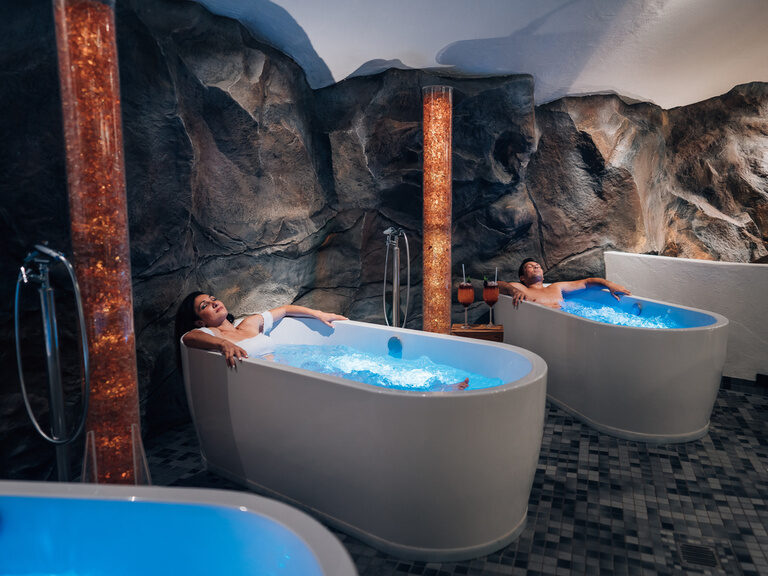 Paar entspannt in zwei separaten Whirlwannen in einer Felsgrotte mit gedämpftem Licht.