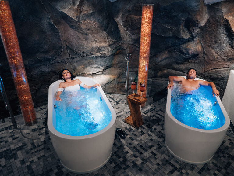 Zwei Personen entspannen in blauen Whirlwannen vor Felswand mit Getränken zwischen ihnen.