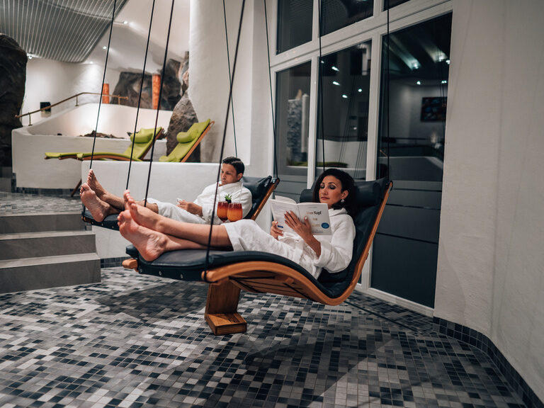 Frau liest im Schwebeliege-Bereich, Mann daneben mit Getränk, beide in Bademänteln.