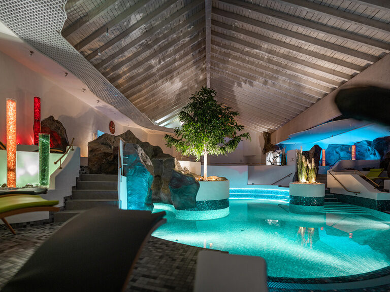 Beleuchteter Indoor-Pool mit Felsen, Liegemöbeln und Lichtskulpturen unter Holzdecke.