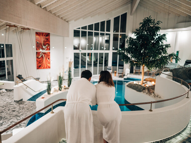 Paar lehnt sich über Geländer mit Blick auf Spa-Bereich mit Pool und Bauminsel.