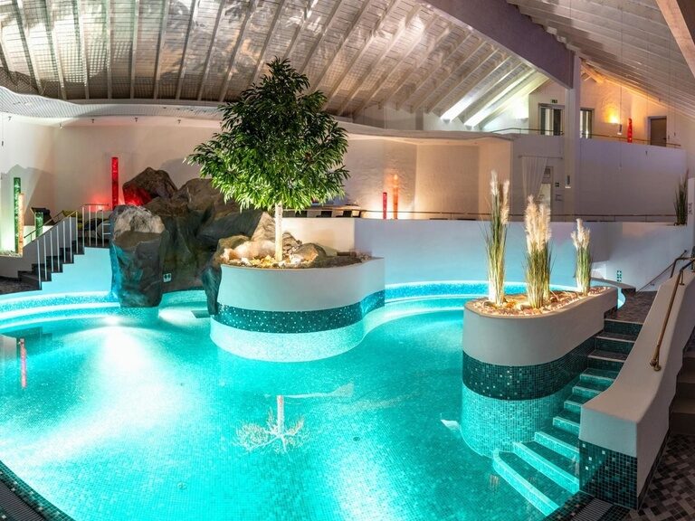 Innenpool mit Wasserinsel, Glasdach und Naturdeko in großzügiger Wellnesslandschaft.