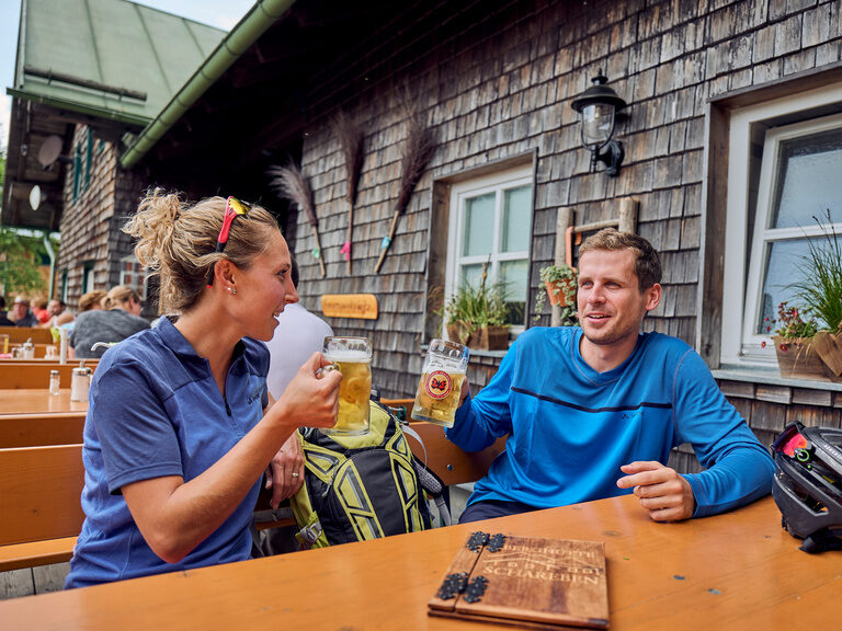 Paar stößt mit Biergläsern im Biergarten an, Fahrradrucksäcke auf der Bank.