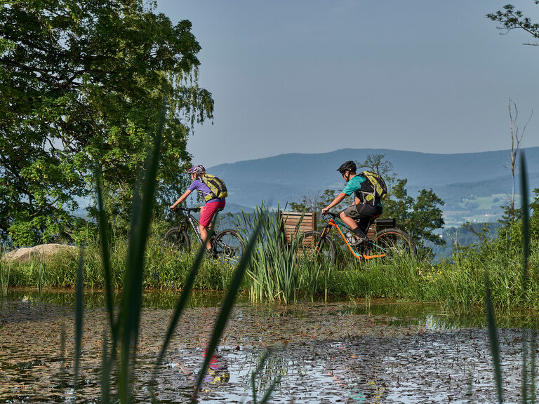 Zwei Mountainbiker fahren am Teich vorbei mit Blick auf hügelige Landschaft.