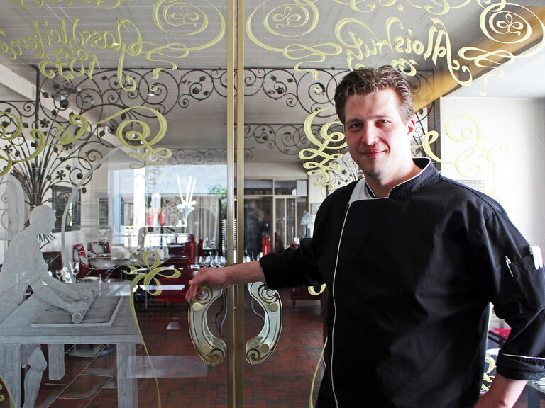 Koch öffnet verglaste Tür mit goldener Verzierung zu einem Restaurant.