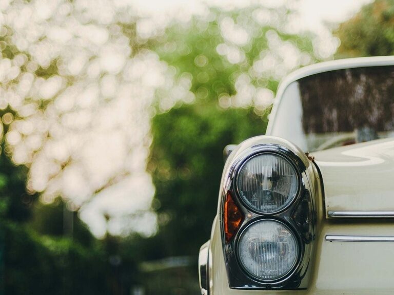 Vorderes Scheinwerferpaar eines Oldtimers vor verschwommenem Waldhintergrund.