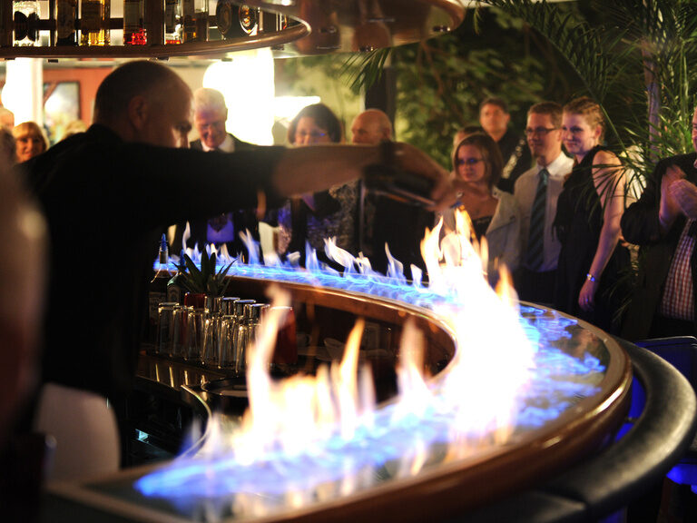 Showbarkeeper entzündet Flammen auf beleuchtetem Tresen vor staunendem Publikum.