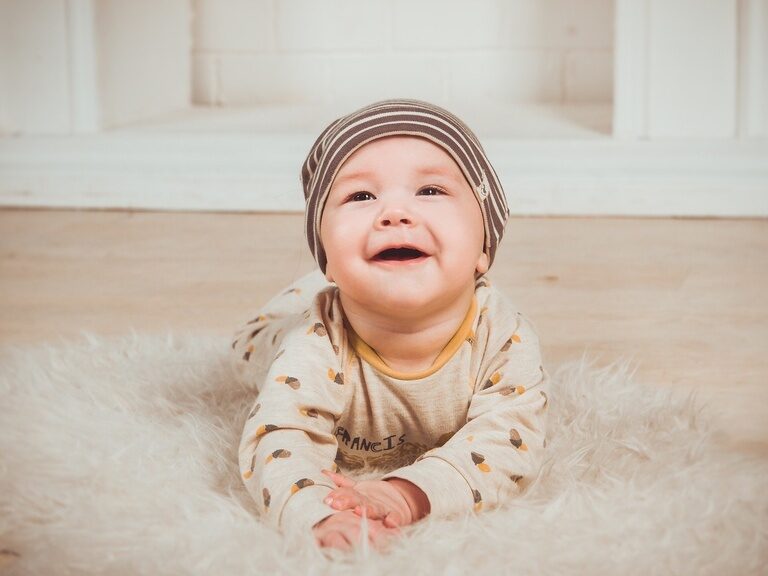 Lachendes Baby mit gestreifter Mütze auf flauschigem Teppich liegend.