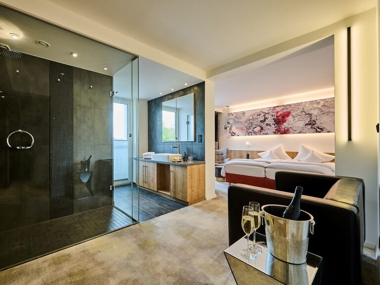 Großzügiges Hotelzimmer mit Glasdusche, Sitzecke und indirekter Beleuchtung.