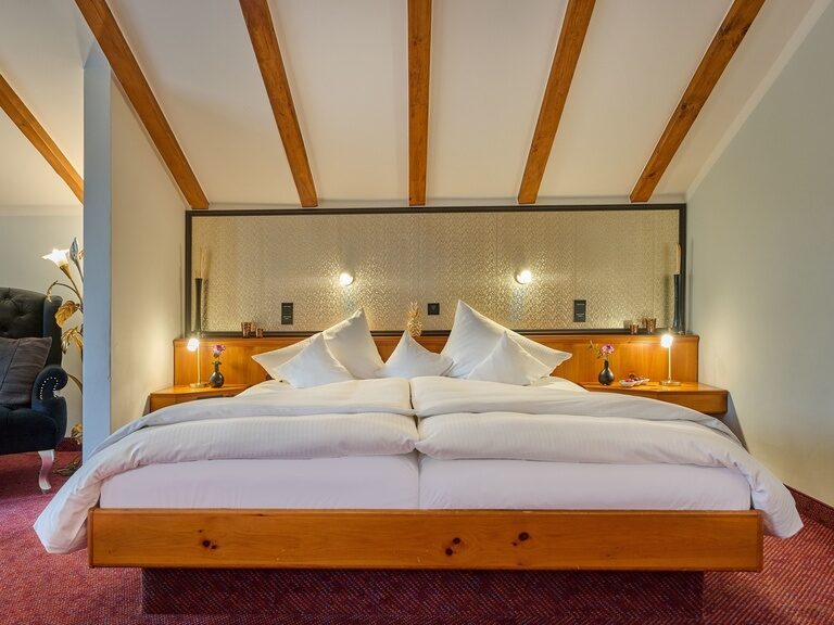 Bettansicht mit Holzbalkendecke, symmetrischer Beleuchtung und grauem Polsterstuhl.