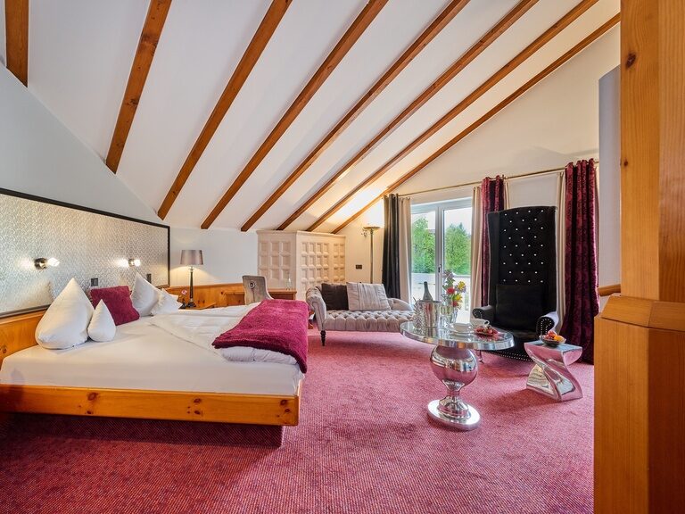 Zimmer mit Doppelbett, edlem Polstersessel, silbernem Tisch und Dachbalken.