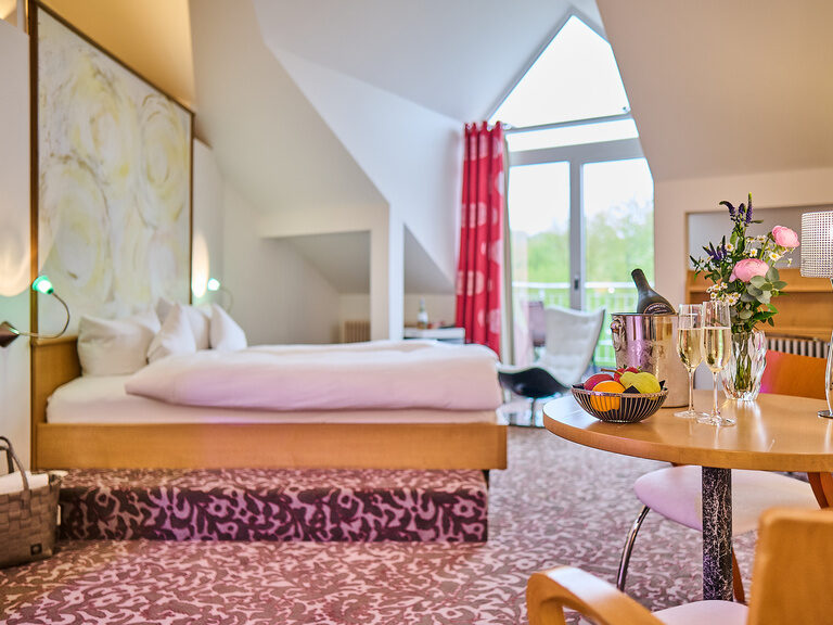 Dachzimmer mit Doppelbett, Tisch mit Blumen und großer Fensterfront.