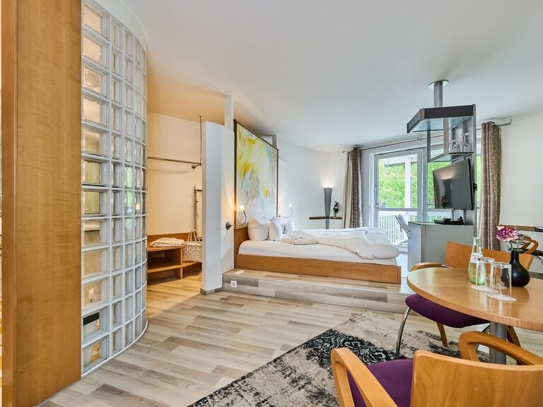 Moderne Dachzimmer-Suite mit Glasblock-Badwand, erhöhtem Bett, Esstisch, Stuhl und Balkon.