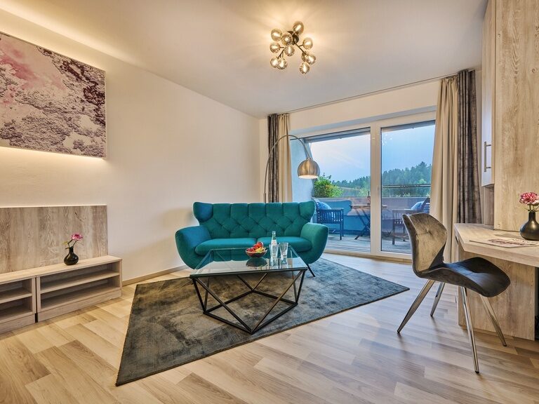 Gemütlicher Wohnbereich im Hotelzimmer mit türkisem Sofa und Balkonzugang.