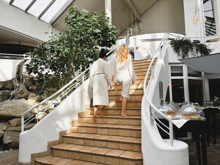Zwei Frauen in Bademänteln steigen eine helle Treppe im Spa-Bereich hinauf.