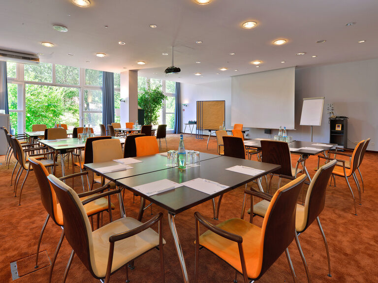 Heller Tagungsraum mit Stuhlreihen, Flipchart, Beamer und bodentiefen Fenstern zur Natur.