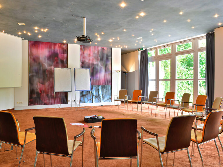 Stuhlkreis in hellem Seminarraum mit Flipcharts und Projektor unter Deckenbeleuchtung.