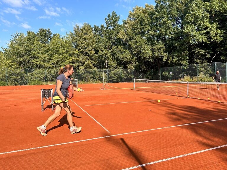 Frau beim Tennis auf Sandplatz mit Ballkorb im Sonnenschein.