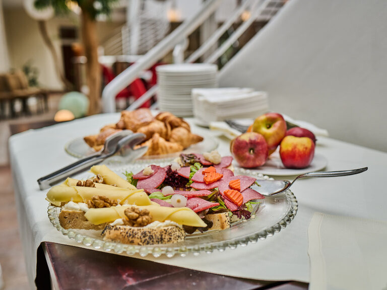 Frühstücksbuffet mit Käse, Wurst, Croissants und Äpfeln auf weißer Tischdecke.