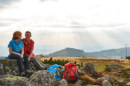 Wanderpaar sitzt auf Felsen mit Rucksäcken, Blick auf bewaldete Mittelgebirgslandschaft.