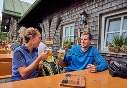 Paar stößt mit Biergläsern im Biergarten an, Fahrradrucksäcke auf der Bank.