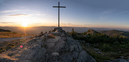 Sonnenaufgang am Gipfelkreuz mit Panoramablick auf bewaldete Berge.